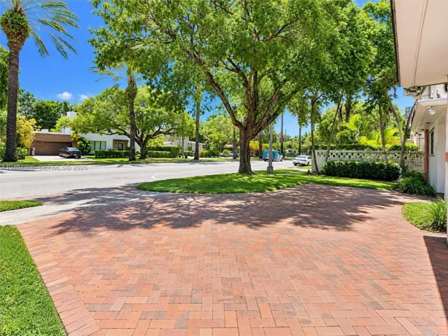 Brick-paver driveway with a herringbone pattern, mature trees, manicured lawn, and perimeter landscaping