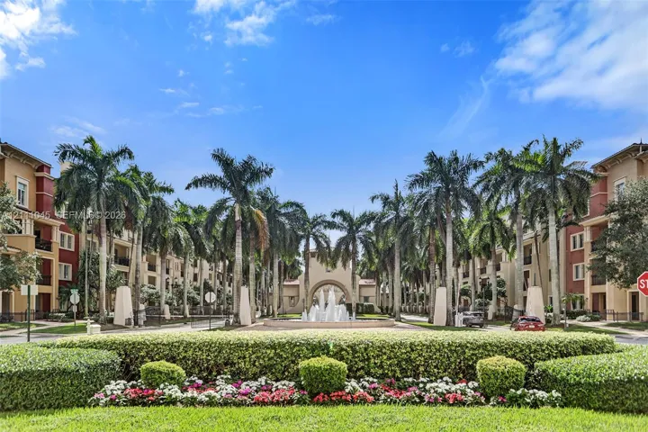 Multi-story residential complex featuring palm-lined drives, manicured hedges, colorful floral beds, and a central tiered water fountain