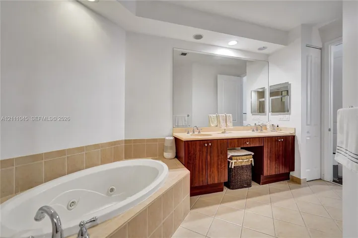 Double vanity with wood-finish cabinetry, solid surface countertop, soaking tub with tile surround, and full-width wall mirror