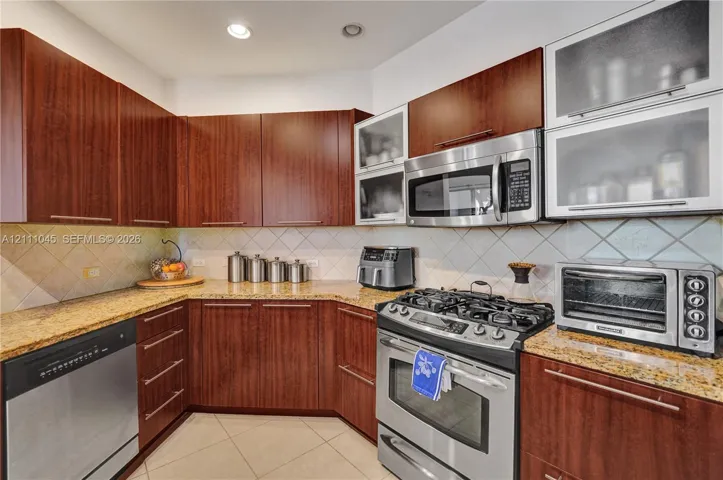 Kitchen featuring warm-tone cabinetry, natural stone countertops, diagonal tile backsplash, stainless steel appliances, and glass-front upper cabinets