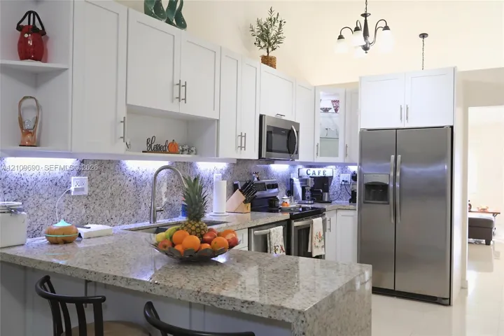 Kitchen featuring white shaker-style cabinetry, stone countertops, stainless steel appliances, under-cabinet lighting, and a multi-arm chandelier