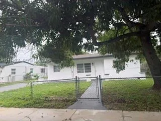 Exterior facade featuring a white painted masonry finish, metal-frame windows, a paved walkway, and a chain-link perimeter fence