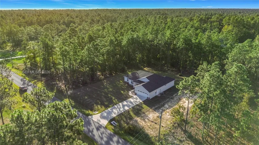 Single-story residence featuring a dark shingled roof, light-toned siding, attached garage, and a paved concrete driveway situated on a wooded lot
