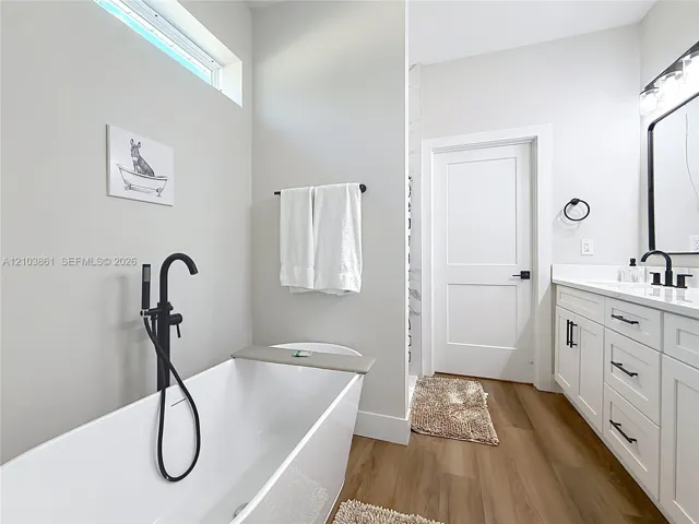 Freestanding soaking tub with a matte black floor-mounted faucet, wood-finish flooring, white shaker-style vanity cabinet with integrated countertop, and transom window