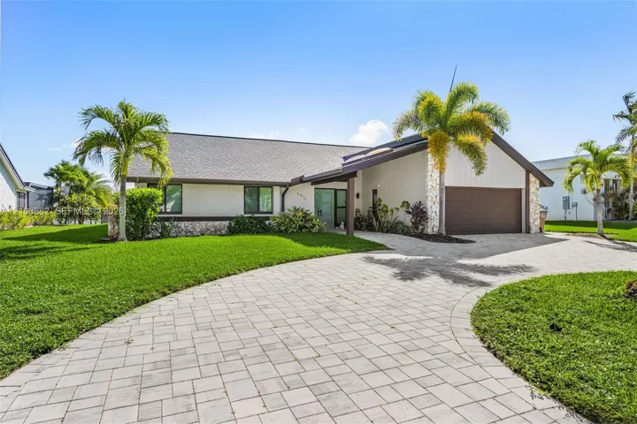 Single-story exterior featuring a stone-clad facade, paver driveway, gabled roofline, integrated garage, and tropical landscaping