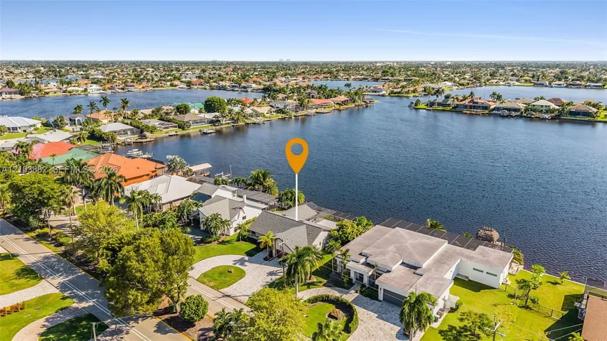 Aerial perspective featuring expansive water frontage, paved circular driveway, mature palms, and residential roofline