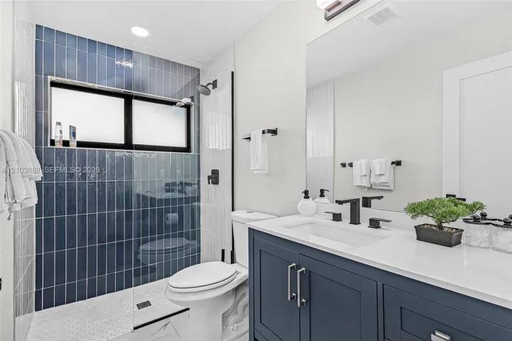 Bathroom featuring a navy blue vanity with a white countertop, wall-to-wall mirror, glass-enclosed shower with blue vertical tile surround, and matte black fixtures