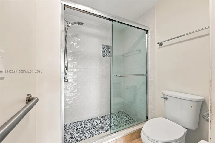 Walk-in shower featuring a sliding glass door, white subway-style wall tiling, patterned mosaic floor tile, and a handheld showerhead