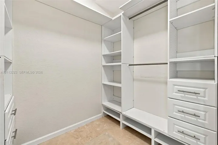 Walk-in closet featuring white built-in shelving, integrated hanging rods, three tiered drawers with metallic pulls, and stone-finish flooring