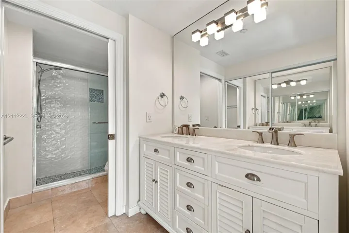 Dual-sink vanity with white cabinetry, stone-surface countertop, walk-in shower with glass enclosure, and wall-mounted light fixture