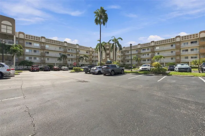 Multi-story residential building featuring covered balconies, stucco facade, surface-level parking, and mature palm trees