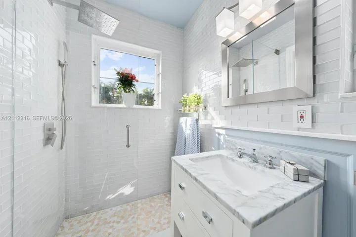 Bathroom featuring a white vanity with a stone countertop, a glass-enclosed shower with pebble-stone flooring, subway tile wall cladding, and a framed mirror with integrated lighting