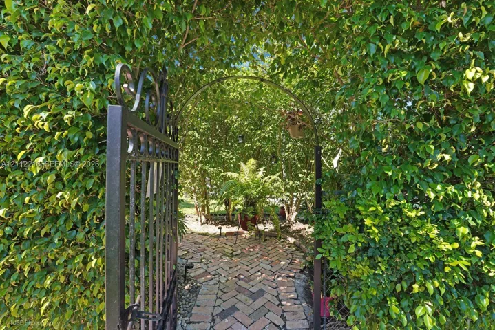 Metal gate with decorative scrollwork, arched entryway, and herringbone-pattern brick pathway lined with mature manicured hedging