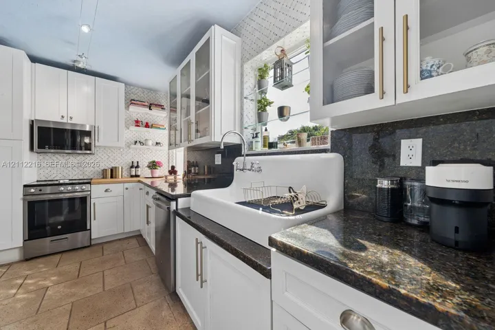 Kitchen featuring white cabinetry with glass-front uppers, apron-front farmhouse sink, stone countertops, stainless steel appliances, and patterned tile backsplash