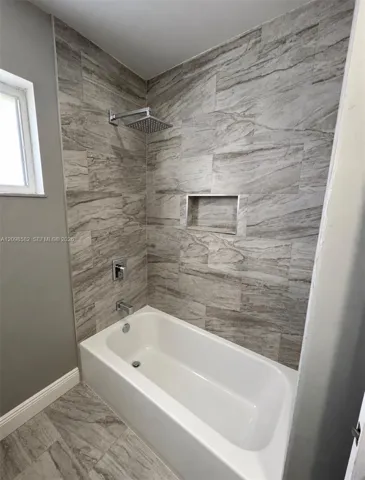 Bathtub and shower combination featuring large-format stone-patterned wall tile, built-in recessed wall niche, stainless steel rainfall showerhead, and rectangular chrome fixtures
