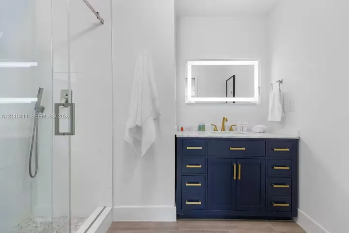 Bathroom featuring a navy blue vanity with brass-finish hardware, stone-surface countertop, frameless glass-enclosed shower with a handheld fixture, and an integrated LED-lighted mirror