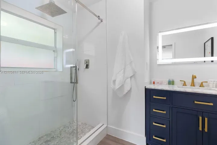 Walk-in shower with a rainfall showerhead, navy blue vanity cabinet with gold-tone hardware, stone-surface countertop, and an integrated LED-lighted mirror