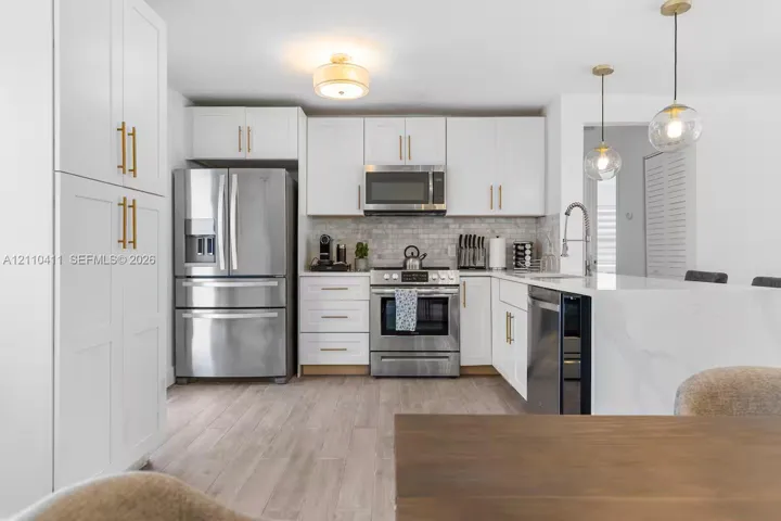 Kitchen featuring white shaker cabinetry with brass hardware, stainless steel appliances, a stone-patterned island, tile backsplash, and wood-finish flooring