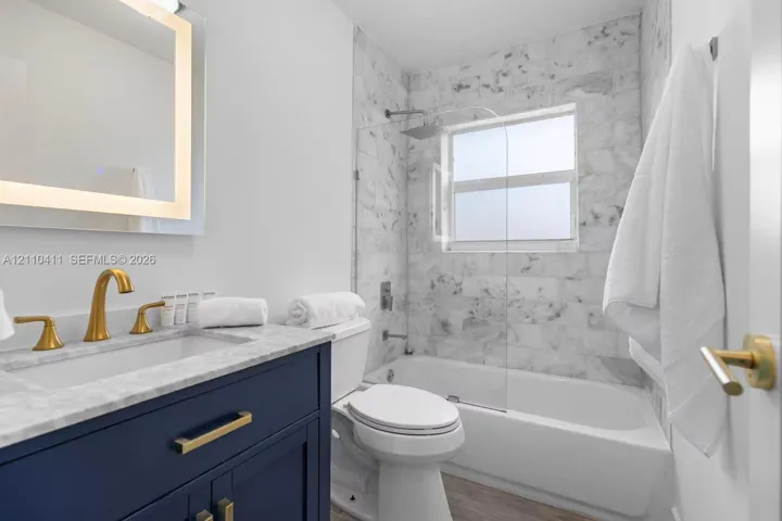 Navy blue vanity with stone-top surface, brushed gold-tone faucet, integrated LED-backlit mirror, stone-tiled shower surround, and glass shower panel