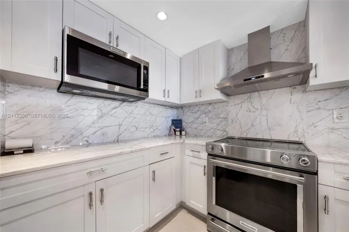 Kitchen featuring white Shaker-style cabinetry, stone-finish countertops, full-height stone-finish backsplash, stainless steel microwave, and stainless steel range with a matching chimney hood