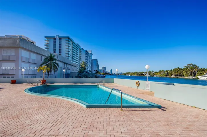 Curved swimming pool featuring a brick-paved deck, perimeter wall, metal handrails, and direct waterfront positioning