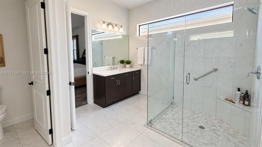 Walk-in shower with stone-finish tile surround, frameless glass enclosure, and built-in bench seating, complemented by a dark wood vanity with white countertop and large vanity mirror