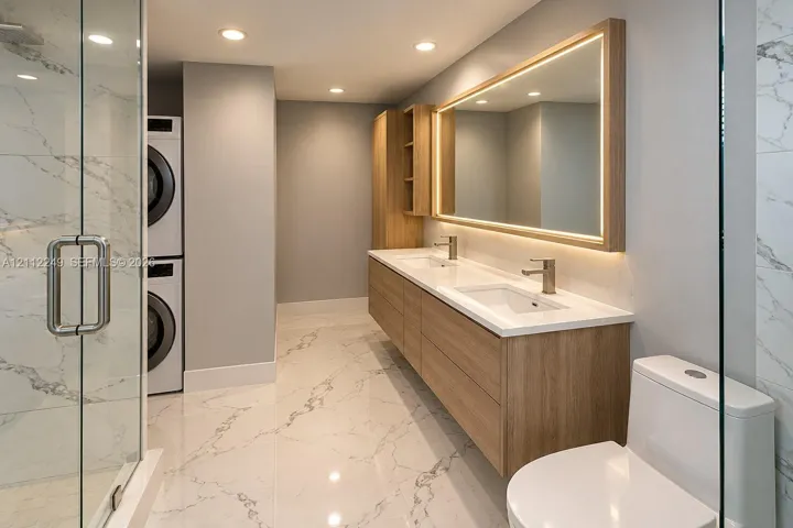 Dual-sink floating vanity with wood-finish cabinetry, integrated LED-backlit mirror, floor-to-ceiling stone-style tile flooring, glass-enclosed shower, and built-in stacked laundry center