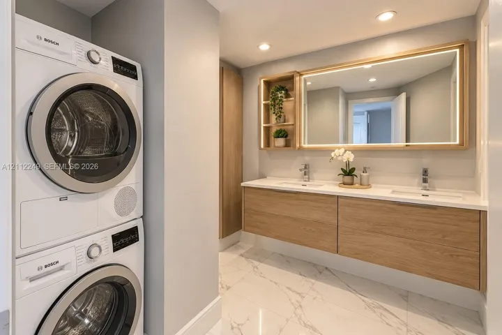 Floating wood-finish vanity with dual integrated sinks, stone-finish countertop, LED-backlit mirror, built-in shelving, and stacked Bosch laundry appliances on polished tile flooring