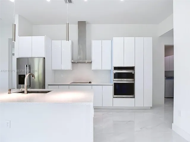 Kitchen featuring white high-gloss cabinetry, stainless steel built-in double ovens and range hood, center island with stone-surface countertop, and large-format porcelain floor tile