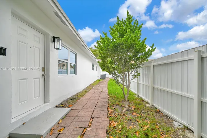 Exterior side path featuring a brick paver walkway, white smooth-stucco siding, a white paneled door, black metal wall-mounted lighting, and a white privacy fence
