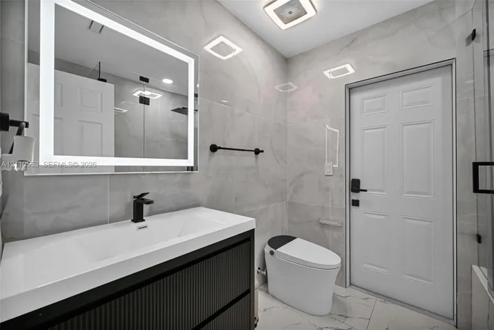 Bathroom featuring a modern vanity with a rectangular integrated sink, wall-mounted matte black faucet, illuminated LED mirror, floor-to-ceiling large-format tile, and a glass-enclosed shower