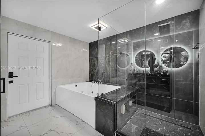 Jetted soaking tub, walk-in shower with glass enclosure, large-format tile flooring, polished stone wall surfacing, and dual illuminated circular mirrors