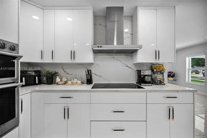 Kitchen featuring glossy white flat-panel cabinetry, white stone-patterned backsplash, stainless steel chimney-style range hood, electric cooktop, and built-in double wall oven