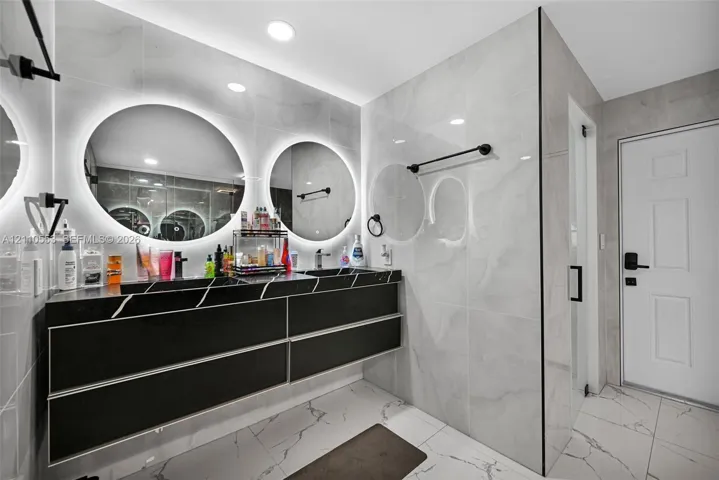 Floating dual-sink vanity with matte black finish, integrated backlit circular mirrors, large-format polished wall tile, and light-toned floor tile with dark veining
