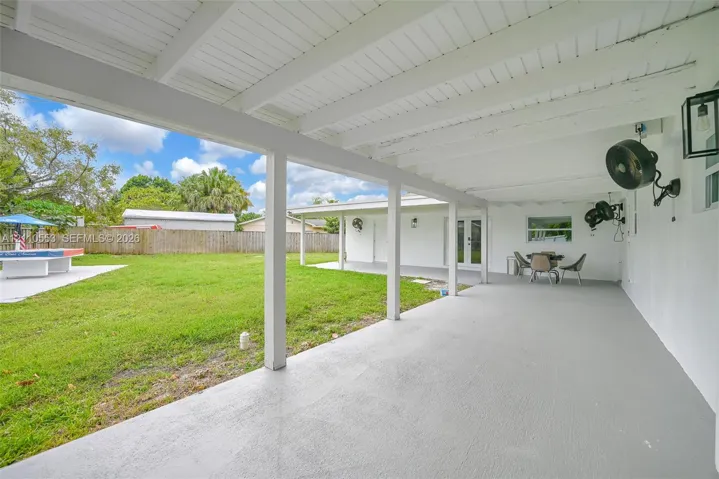 Covered rear patio featuring a painted concrete floor, wood-beam ceiling, wall-mounted outdoor lighting, and expansive backyard space enclosed by wood fencing