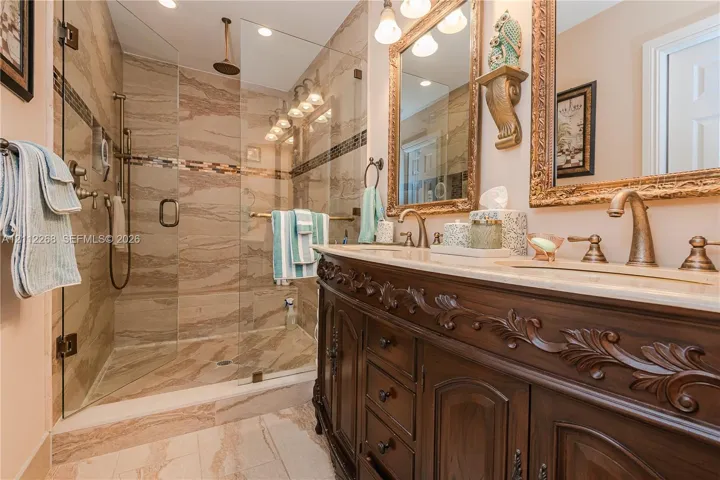 Bathroom featuring an expansive wood vanity with intricate carved detailing, a glass-enclosed walk-in shower with floor-to-ceiling tile surround, stone-finish countertops, dual-sink configuration, and brass-tone plumbing fixtures