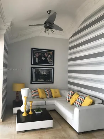 Vaulted ceiling featuring ornate crown molding and a multi-blade ceiling fan with integrated lighting, finished with patterned wall coverings and stone-finish flooring
