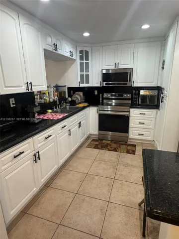 Kitchen featuring white shaker-style cabinetry with matte black hardware, dark stone countertops, stainless steel appliances, glass-panel upper cabinet doors, and tile flooring