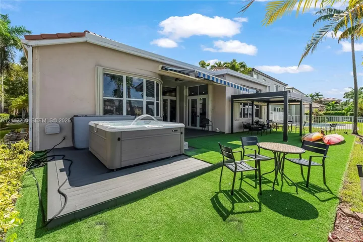 Rear exterior featuring an integrated spa, elevated composite decking, a fixed metal-frame pergola, and artificial turf landscaping