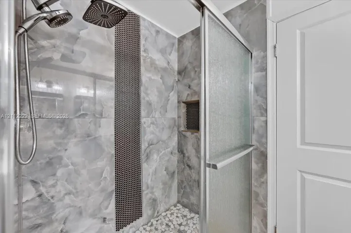 Walk-in shower featuring large-format wall tile with a contrasting mosaic accent strip, rain-style shower head, handheld sprayer, pebble-style shower floor, and a recessed wall niche