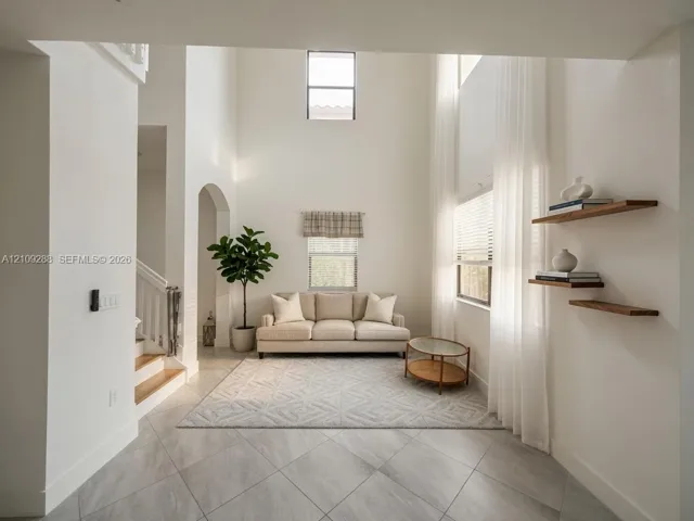 Double-height ceilings featuring natural lighting from multiple window levels, neutral stone-finish tile flooring, arched architectural detailing, and wall-mounted wood-finish floating shelves