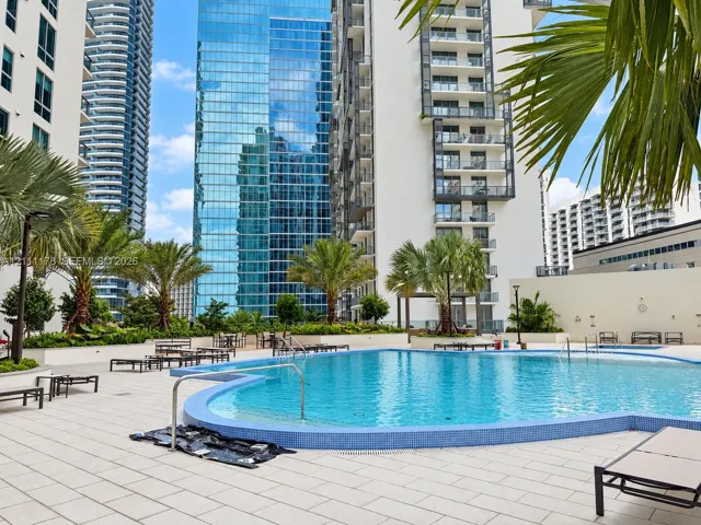 Curved swimming pool featuring tile coping, surrounding paved deck, integrated landscaping, and palm trees