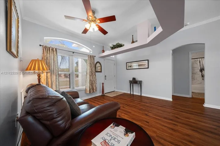 Living area featuring wood-finish flooring, vaulted ceilings with a decorative plant ledge, a ceiling fan with integrated lighting, and an arched window