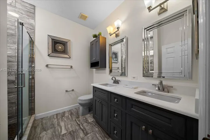 Dual-sink vanity with a solid-surface countertop, dark-finished cabinetry, walk-in glass shower enclosure, wood-finish tile flooring, and wall-mounted light fixtures