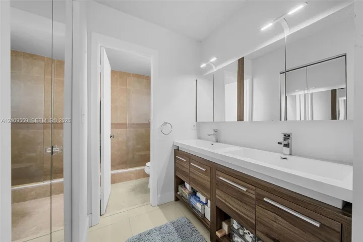 Dual-sink vanity featuring wood-finish cabinetry, integrated rectangular basins, and full-width mirror cabinet storage, complemented by a glass-enclosed shower with tile surround