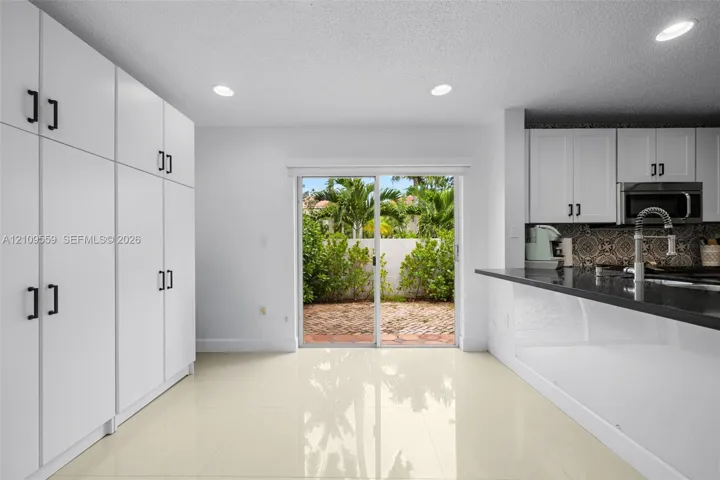 Kitchen featuring crisp white floor-to-ceiling cabinetry, dark stone countertops, patterned tile backsplash, polished neutral tile flooring, and sliding glass doors with exterior brick patio access