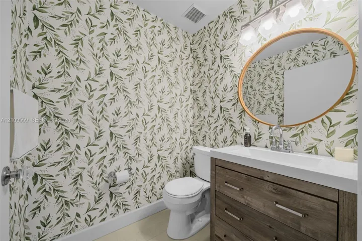 Restroom featuring a wood-finish vanity, white rectangular countertop sink, oval gold-rimmed mirror, and botanical wallpaper