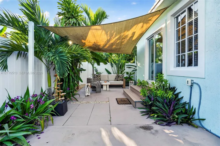 Paved patio area featuring a mounted shade sail, wood-finish stepped platform, light-colored stucco siding, and surrounding privacy fencing