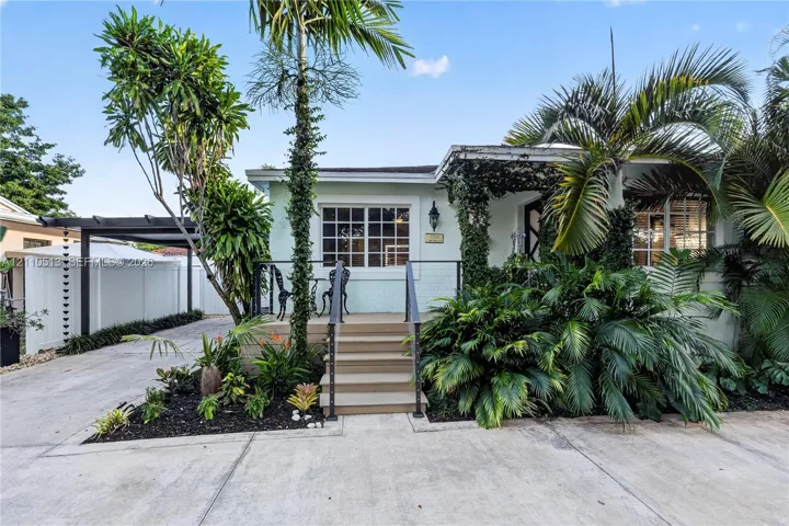 Light-colored exterior siding with grid-pattern windows, raised wood-finish front porch, black metal railing, concrete driveway, and covered carport