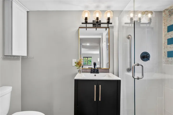 Bathroom featuring a vanity with dark cabinetry and a light-toned countertop, three-light fixture, gold-framed mirror, and a glass-enclosed shower with tiled surround